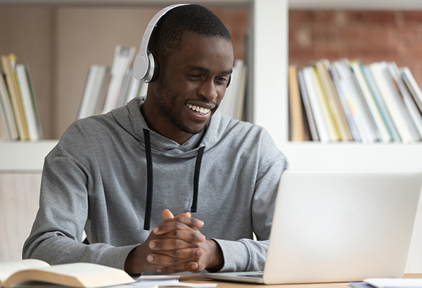 Student with headphones smiles while looking at computer with open book next to him